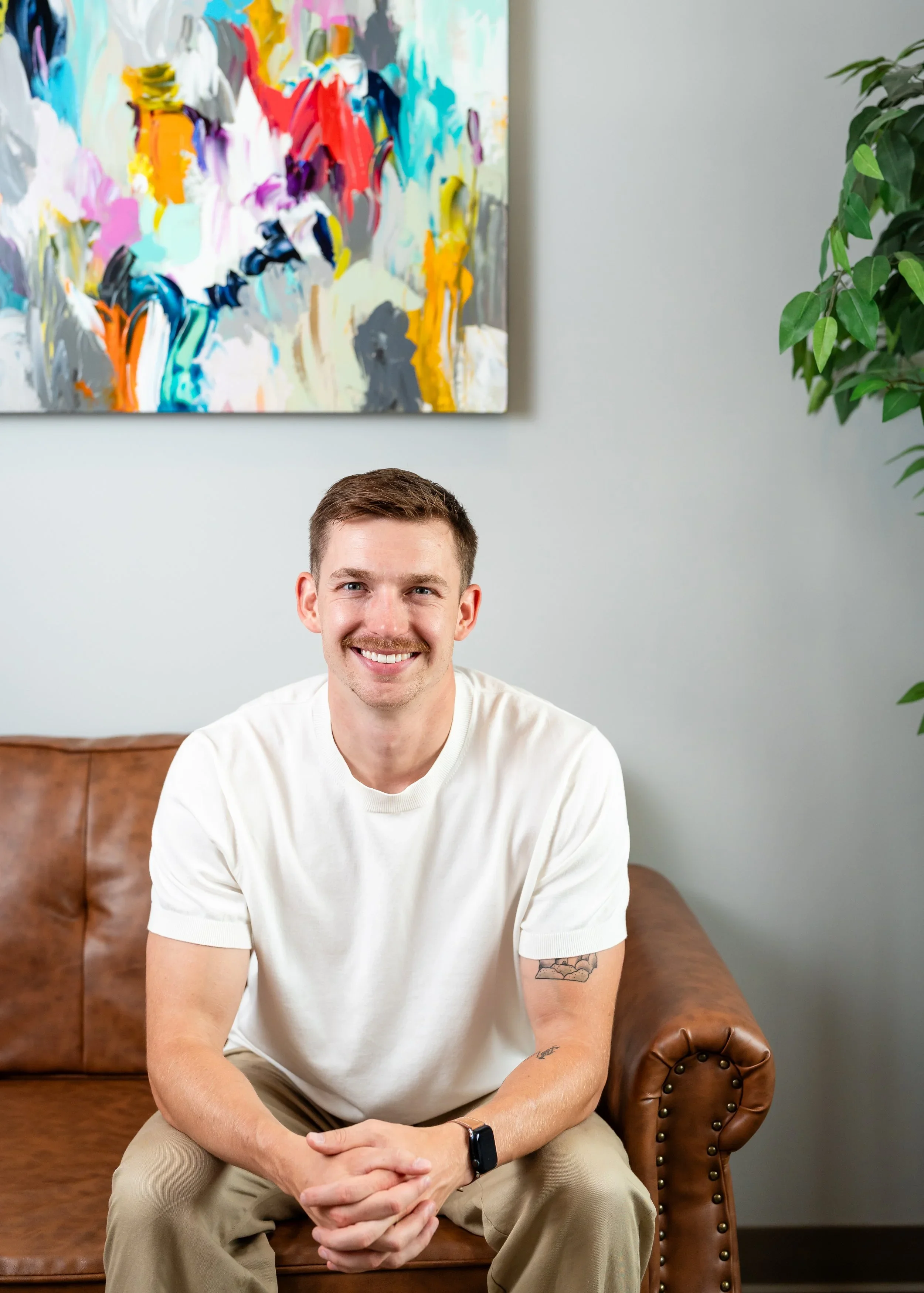A smiling man with short brown hair, a mustache, and a beard sitting on a brown leather couch in a modern room with abstract colorful artwork on the wall and green plants.