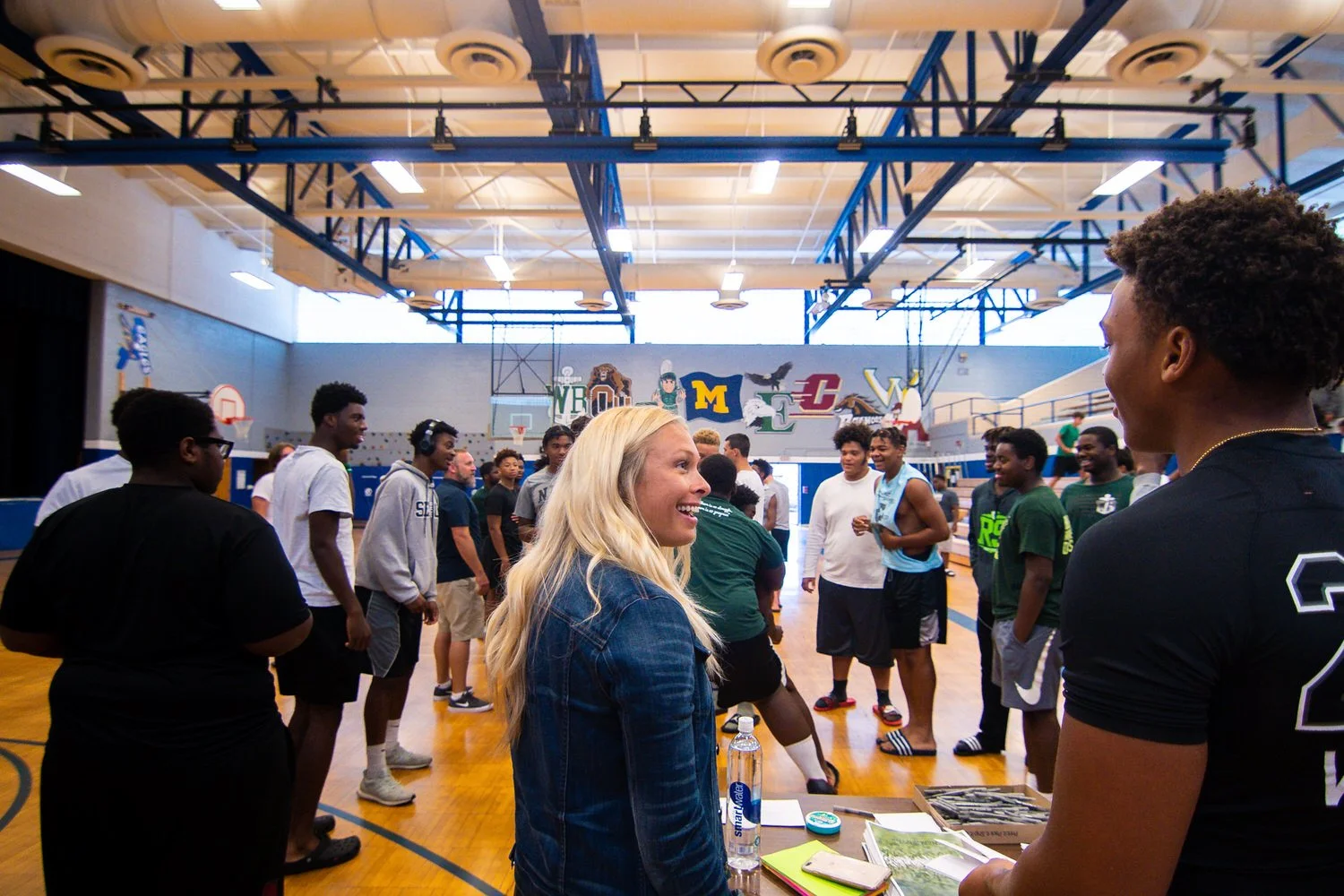 Group of students in a gymnasium, some in athletic attire, chatting and socializing, with a woman with blonde hair smiling at a young man with curly hair.