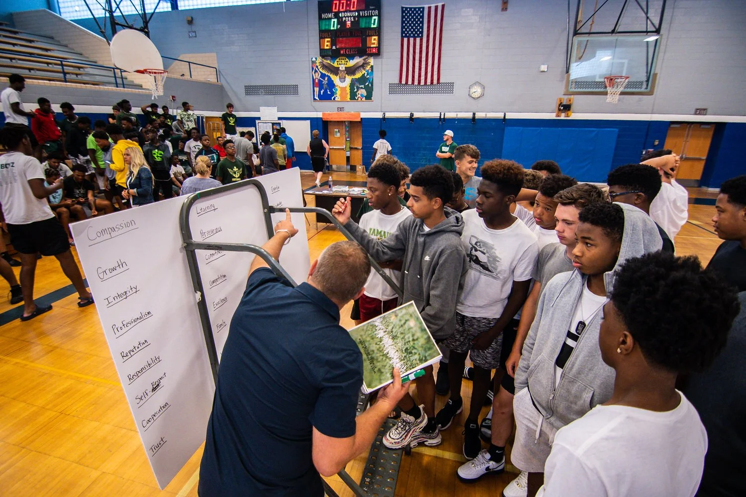 A group of boys listening to a speaker during a workshop in a school gymnasium. The speaker is pointing to a whiteboard with words like 'Compassion,' 'Growth,' 'Integrity,' 'Reputation,' and others written on it. In the background, more students are seated and standing, and the gym features American flags, a scoreboard, and sports equipment.