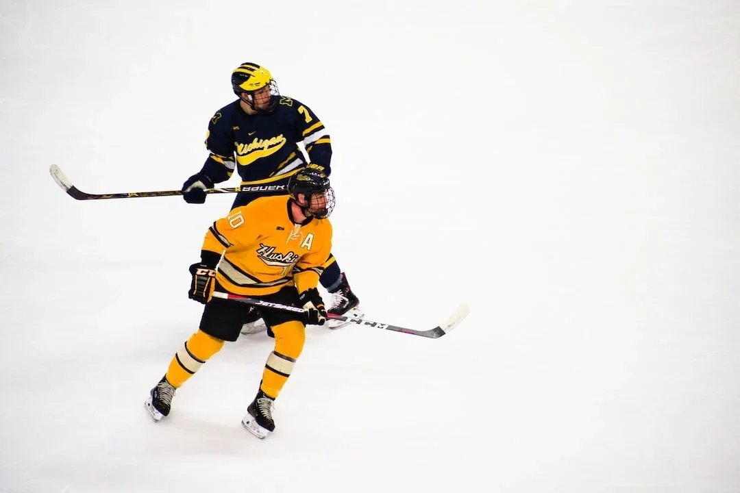 Two ice hockey players in yellow and navy uniforms compete on the ice, with a mostly empty background.
