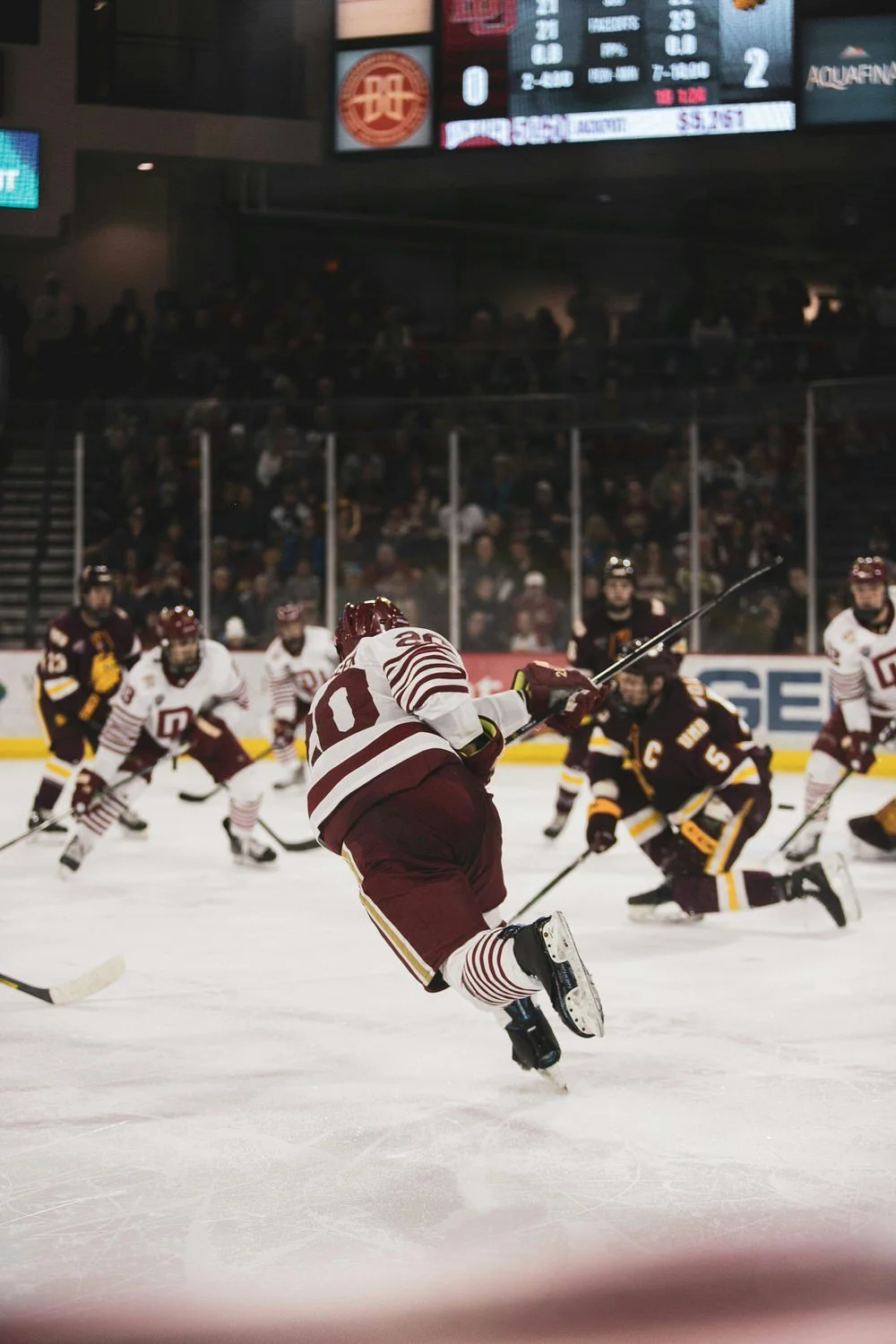 An ice hockey game in progress with players in maroon and white jerseys and black, yellow, and white jerseys on ice, with spectators in the background.