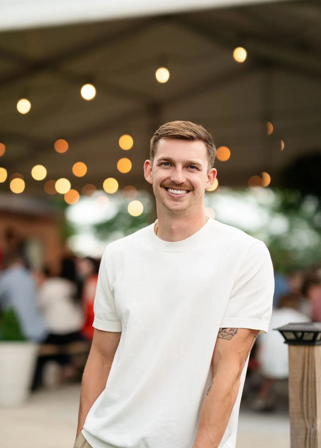 A smiling young man with short brown hair, a mustache, wearing a white t-shirt, standing outdoors at a social gathering with blurred people and string lights in the background.