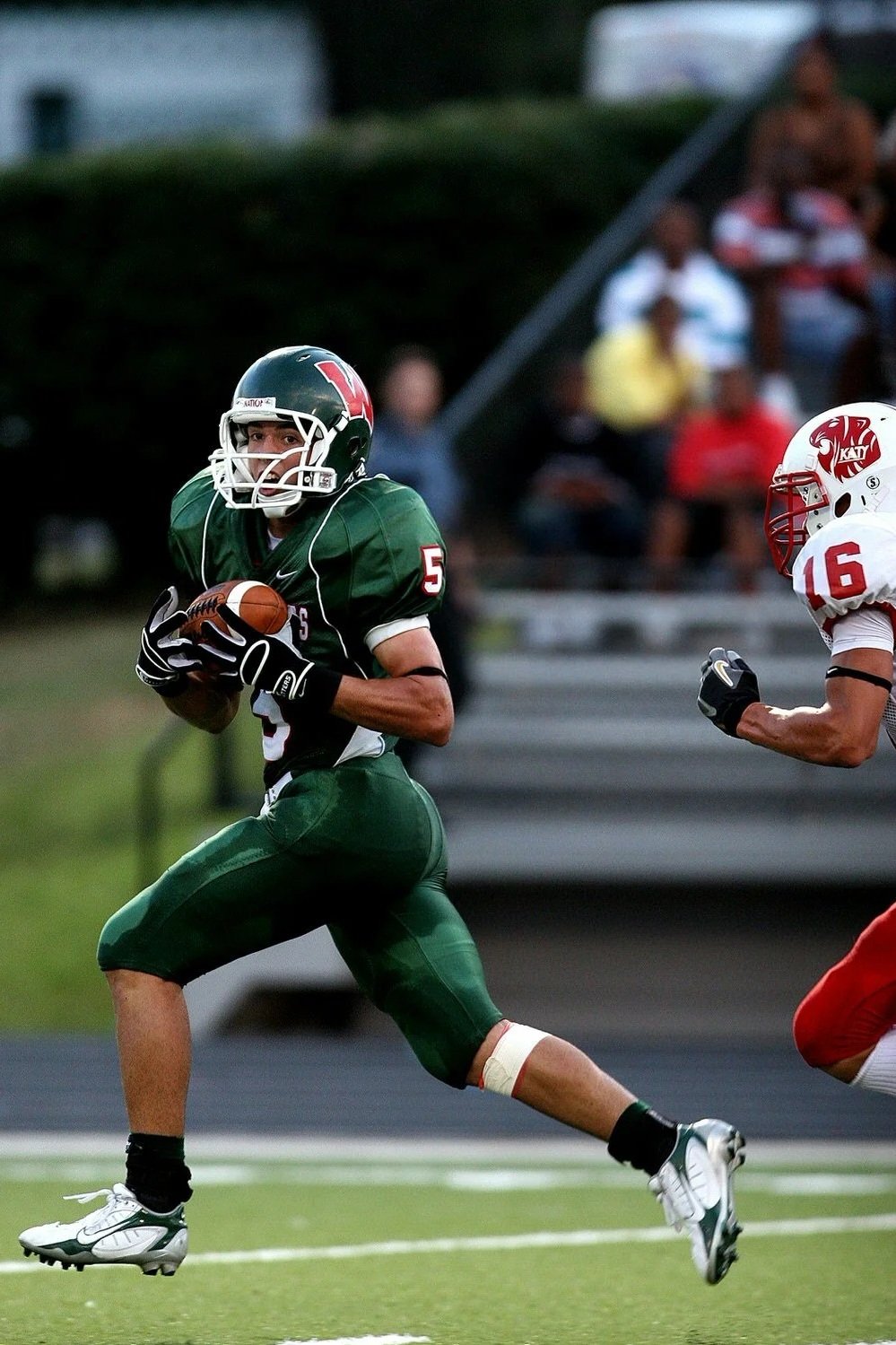 Football player in green uniform running with the ball in a game, with a player in white and red chasing behind, on a field with spectators in the background.