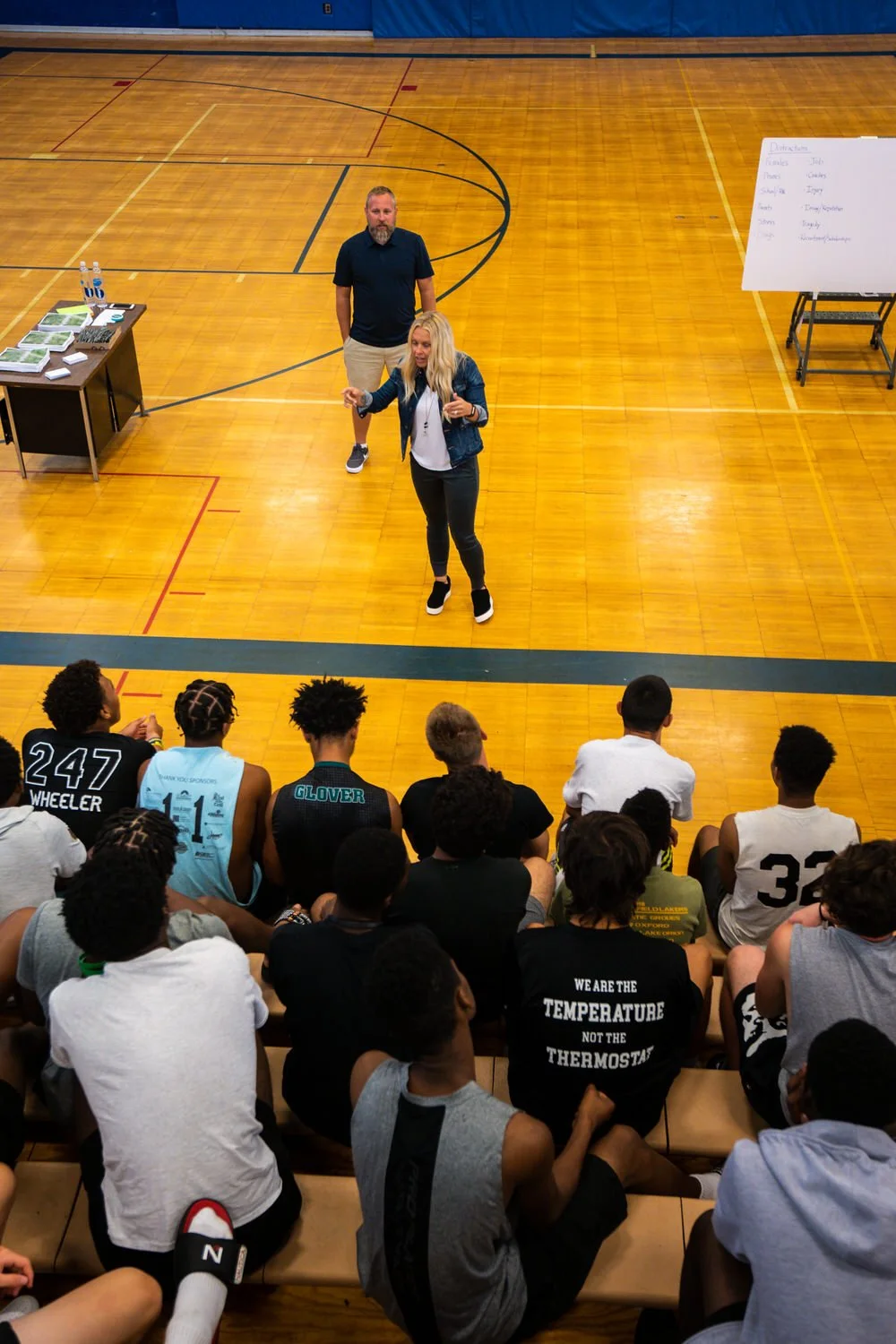 A woman speaking to a group of young basketball players in a gymnasium, with two people standing nearby and a whiteboard in the background.