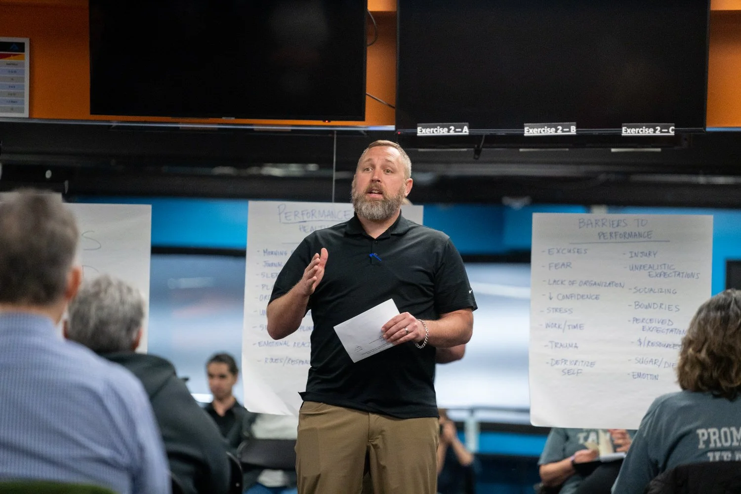 A man with a beard and short hair giving a presentation to a group of people in a room with blue walls, a large black monitor, and posters with handwritten notes.