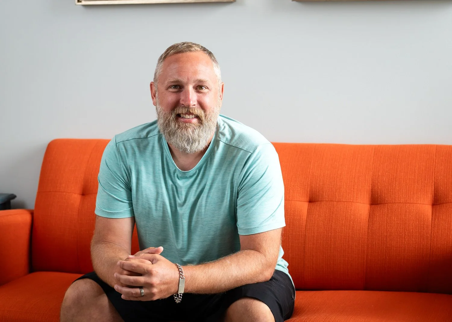 A man with a gray beard and short hair, wearing a light blue t-shirt and black shorts, sitting on an orange couch with a white wall in the background.