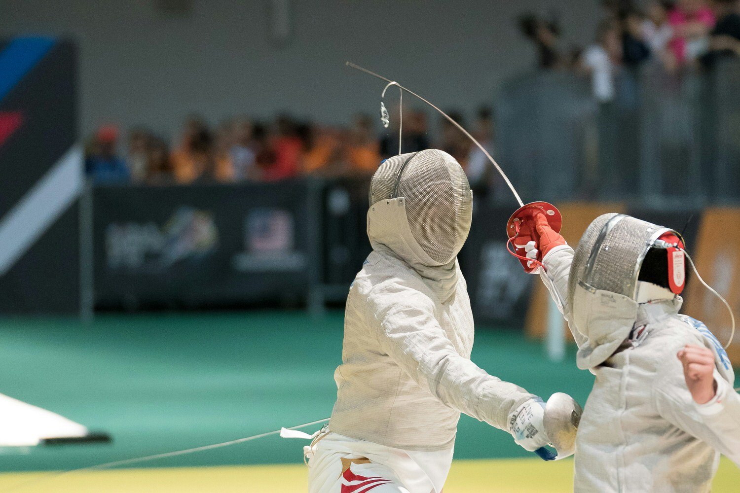 Two fencers in white protective gear and masks, engaged in a fencing match, with one fencer holding their foil in a fencing stance as they approach each other on the fencing piste.