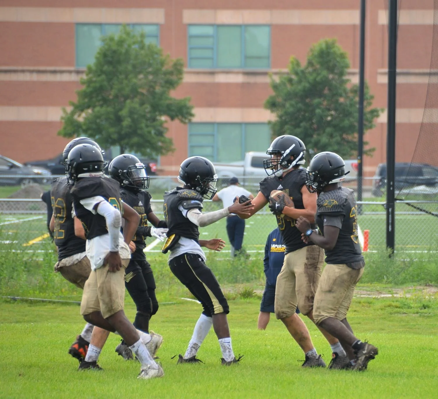 Youth football players on a field during practice, with some wearing black jerseys with yellow and white accents and helmets, passing a football, with a brick building and trees in the background.