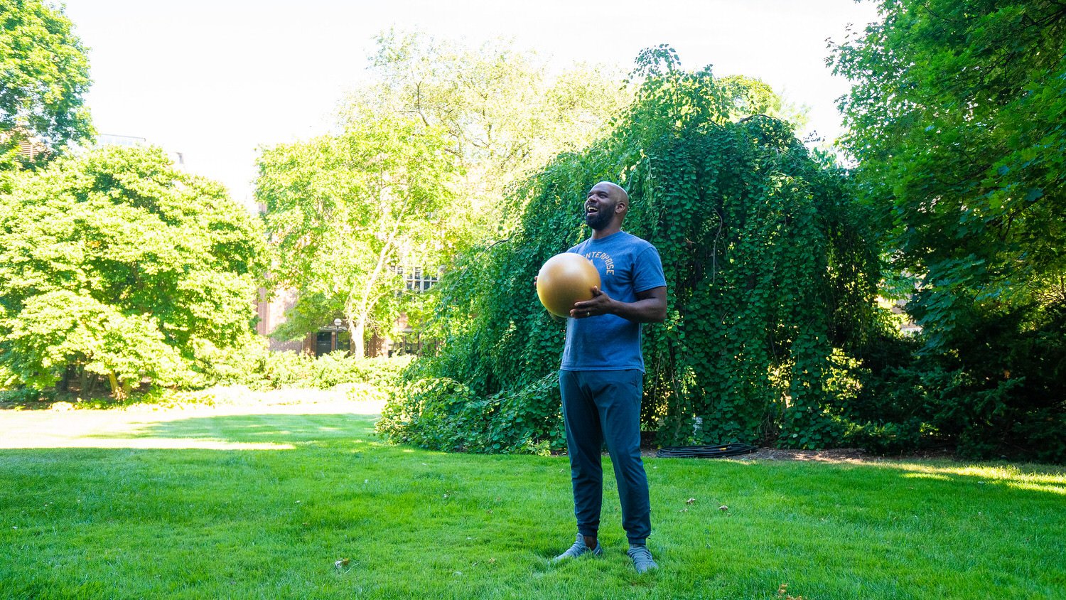 A man in athletic clothing holding a medicine ball in a park with lush green trees and grass.