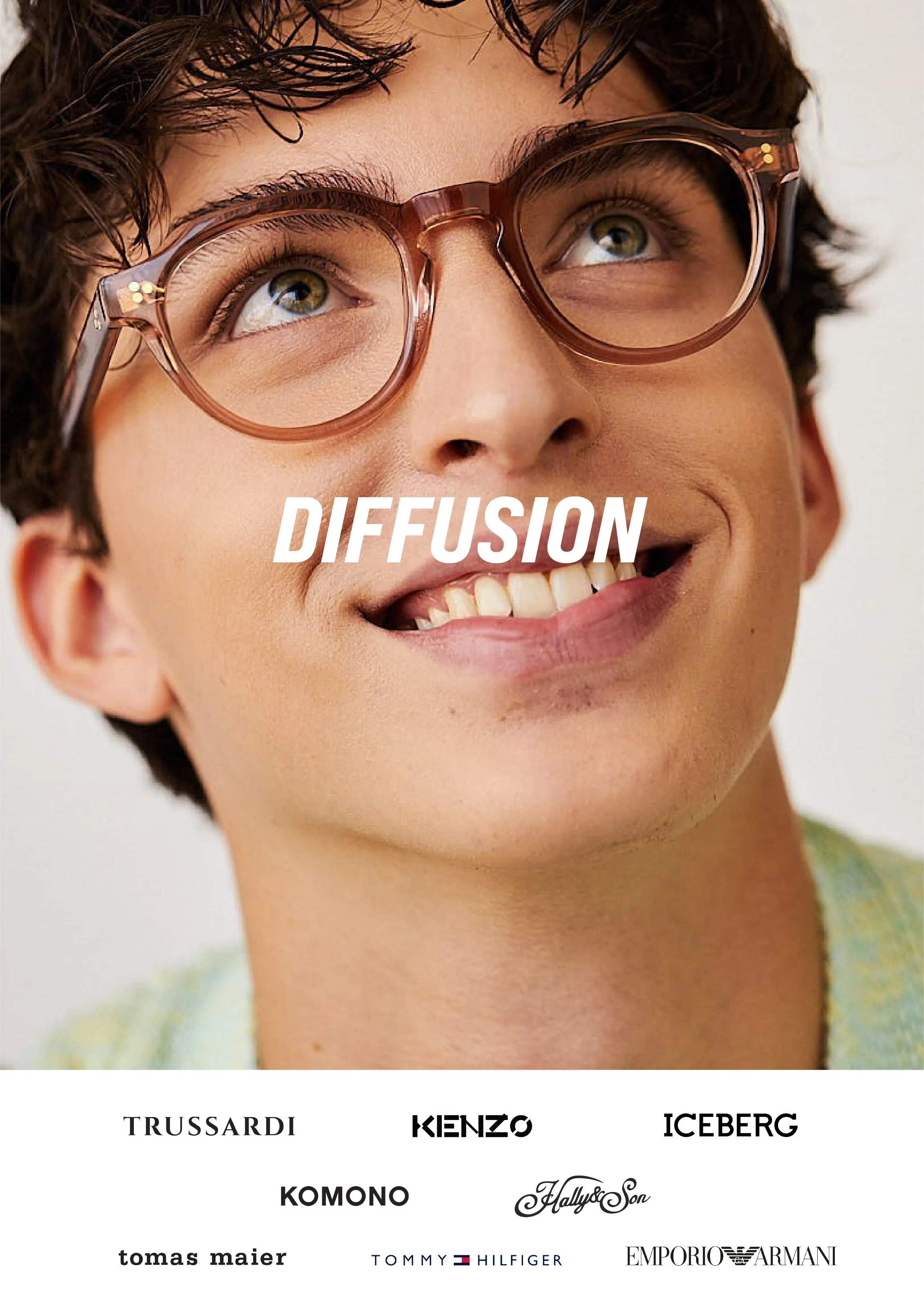 Close-up of a person with short dark hair wearing large brown glasses, smiling with visible teeth, and looking slightly upwards. The background is neutral, and the lower part of the image features brand logos and names such as Trussardi, Kenzo, Iceberg, Komono, Fallys & Son, Tomas Maier, Tommy Hilfiger, and Emporio Armani.