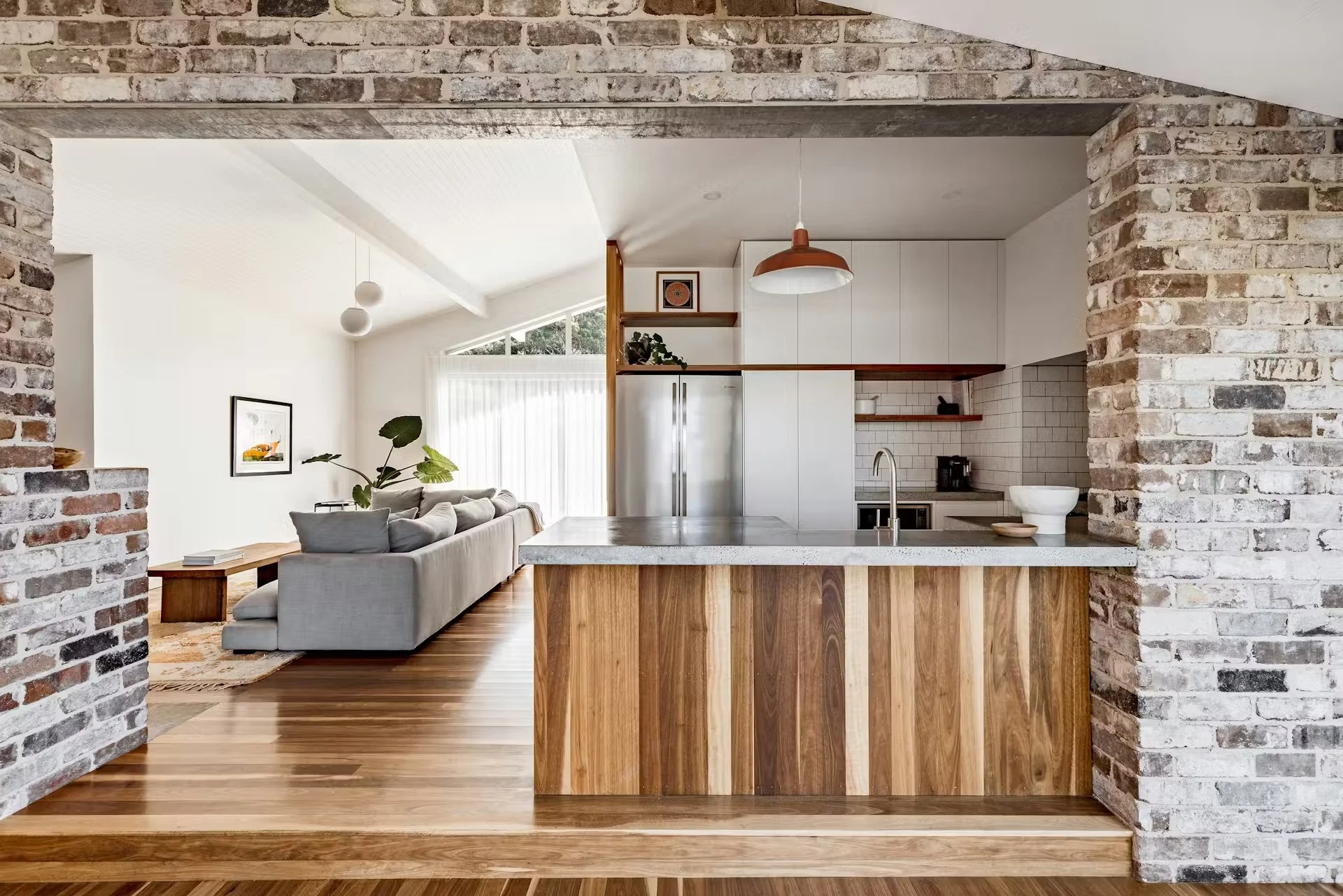 70's Beach House open-concept living room and kitchen with brick archway, white walls, timber flooring, and natural light from large windows.