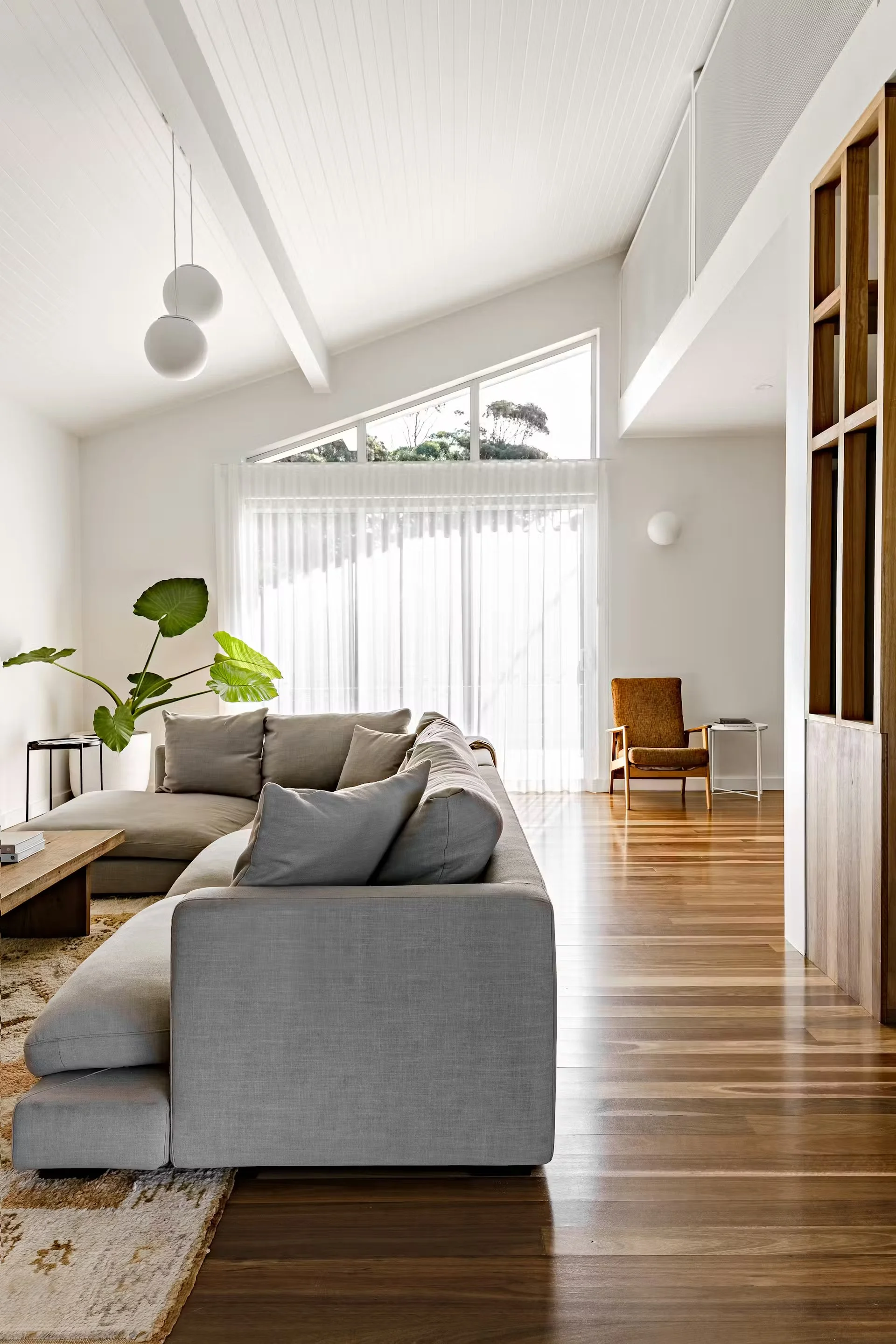 70's Beach House interiors: A bright living room with a high, angled ceiling, large windows, and timber flooring.