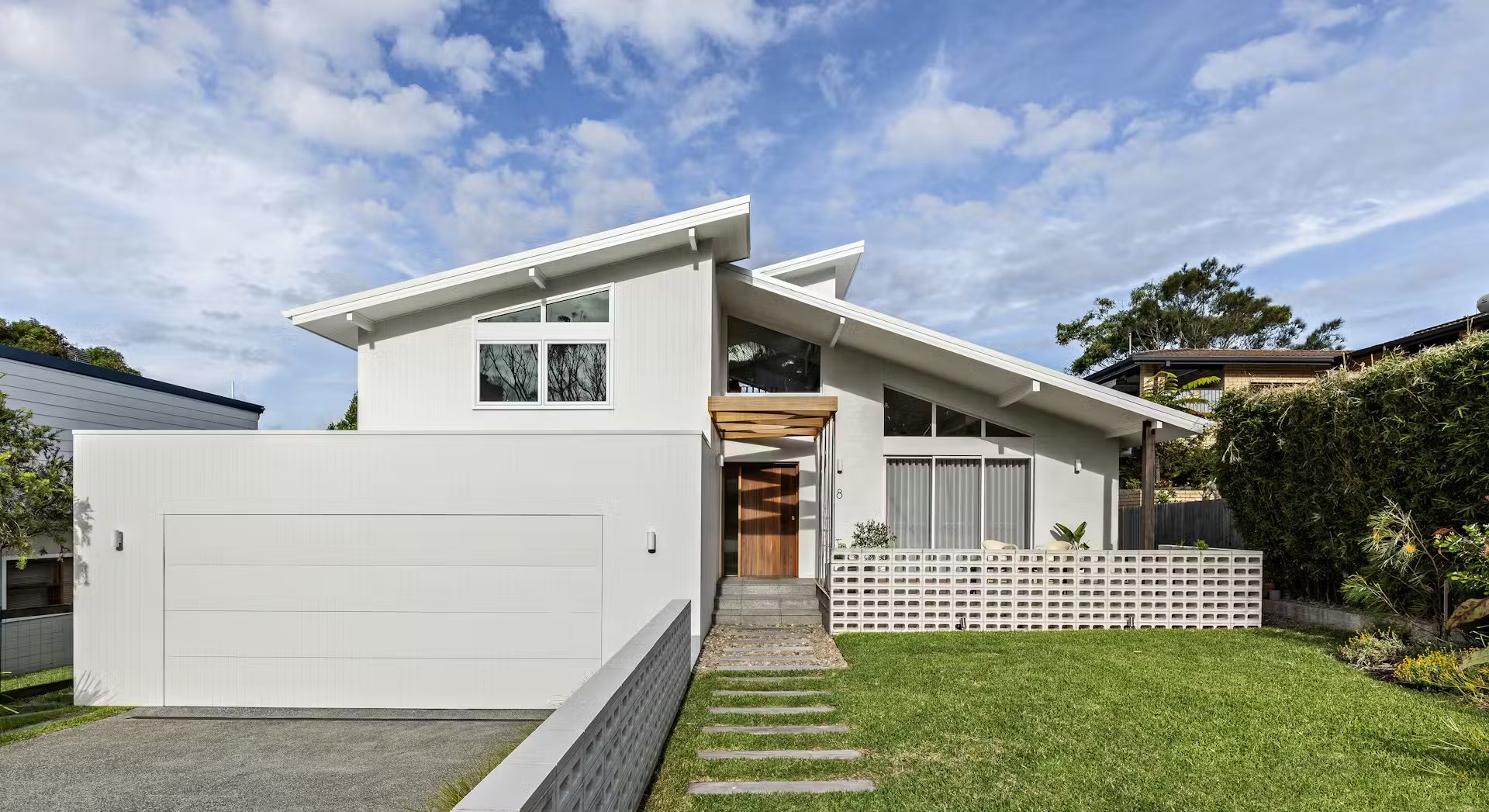 Modern white house with a unique sloped roof, wooden door, and large windows, surrounded by greenery under a partly cloudy sky.