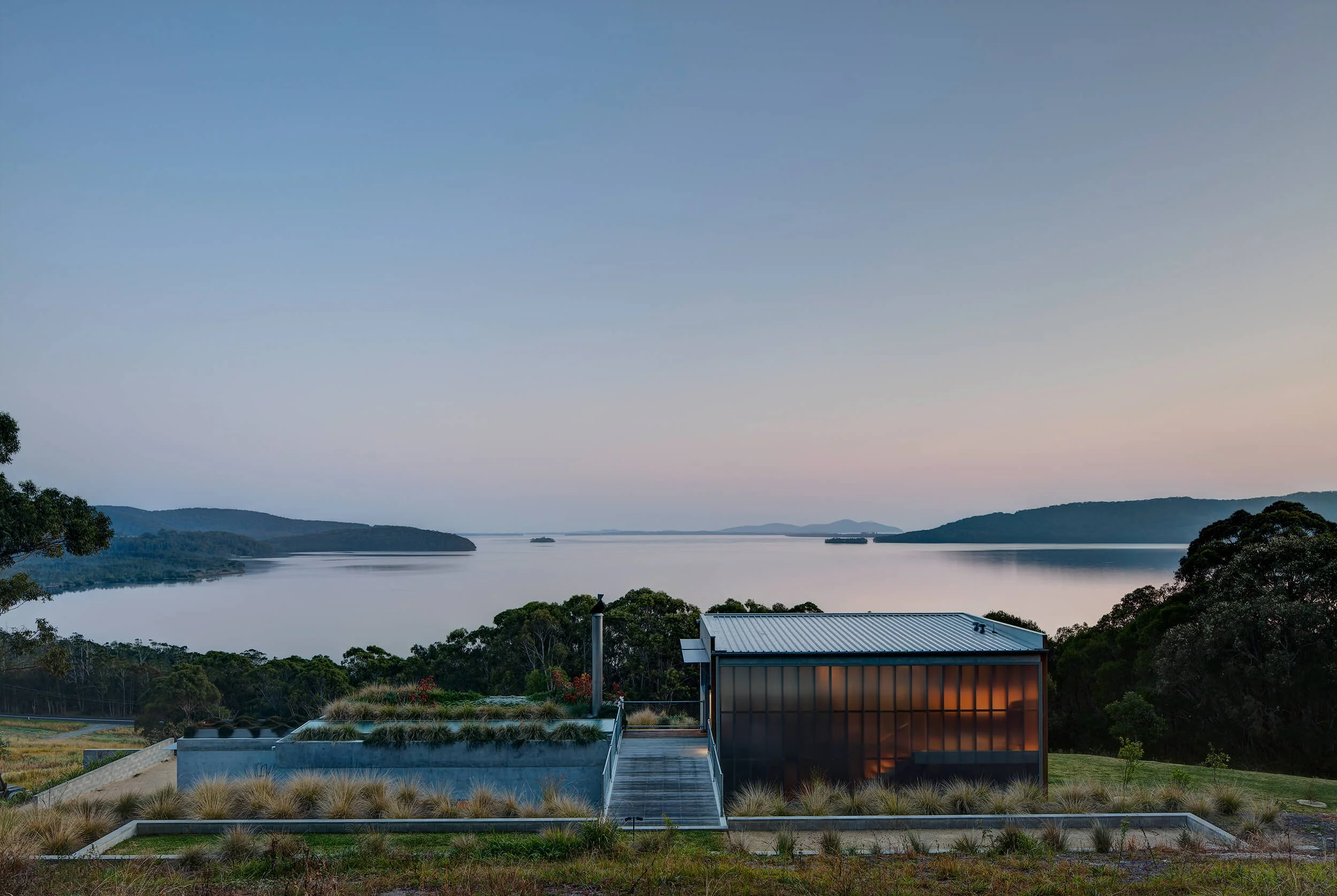 A lakeside view with mountains in the background, a calm water body, and a modern building with a metal roof in the foreground.