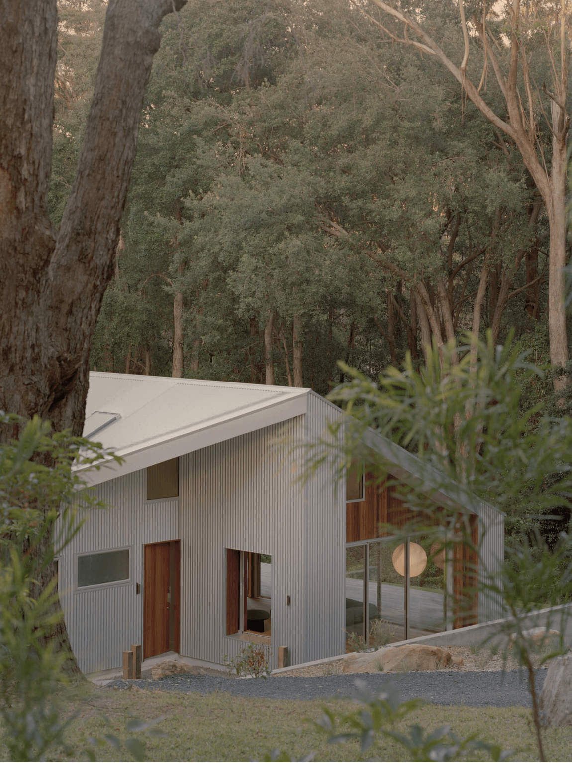 Clad in corrugated Zincalume steel and a white gabled roof, surrounded by trees and greenery, with a view through the foliage.