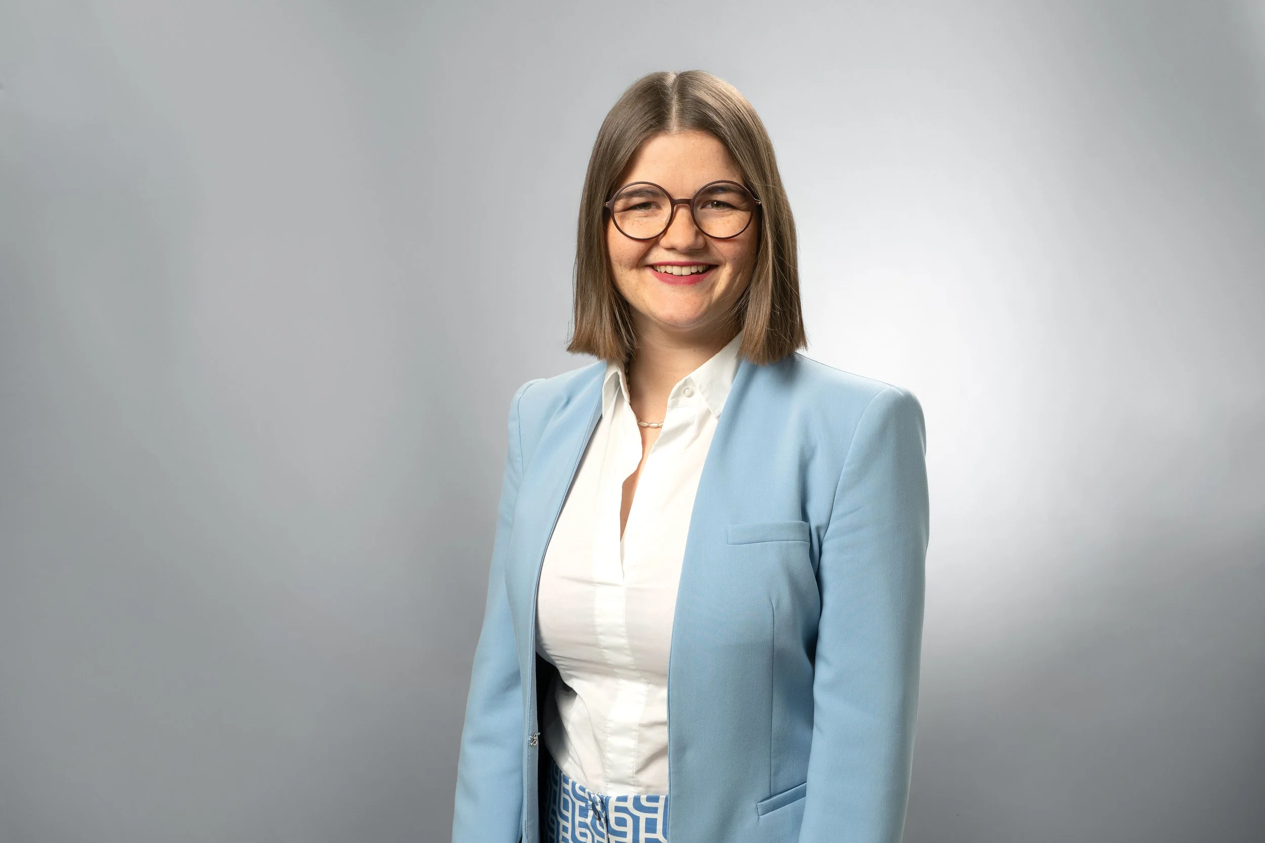 A young woman with shoulder-length brown hair and glasses, wearing a light blue blazer over a white blouse, smiling against a gray background.