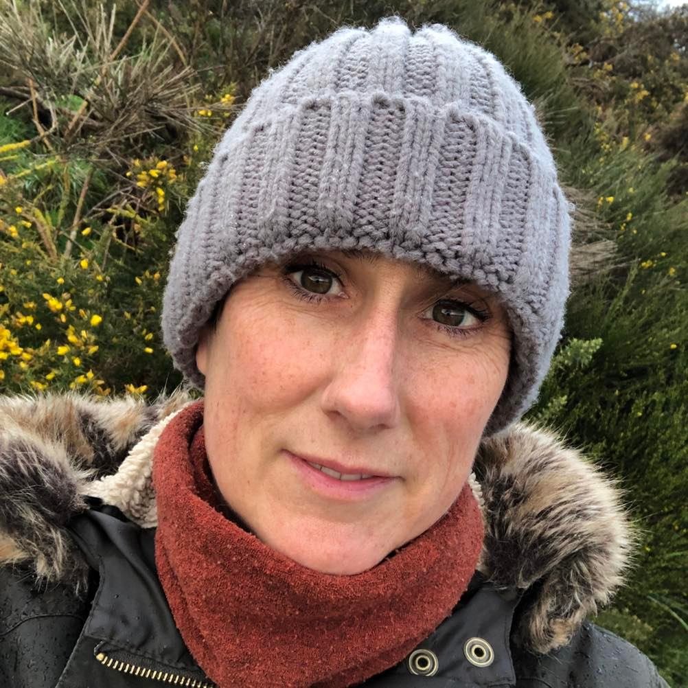 A woman wearing a gray knit beanie, a red scarf, and a black winter coat with a faux fur-lined hood, outdoors in front of green bushes with yellow flowers.