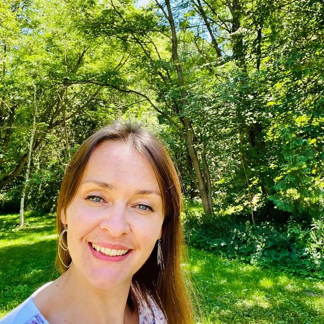 Woman smiling outdoors in a green, wooded park on a sunny day.