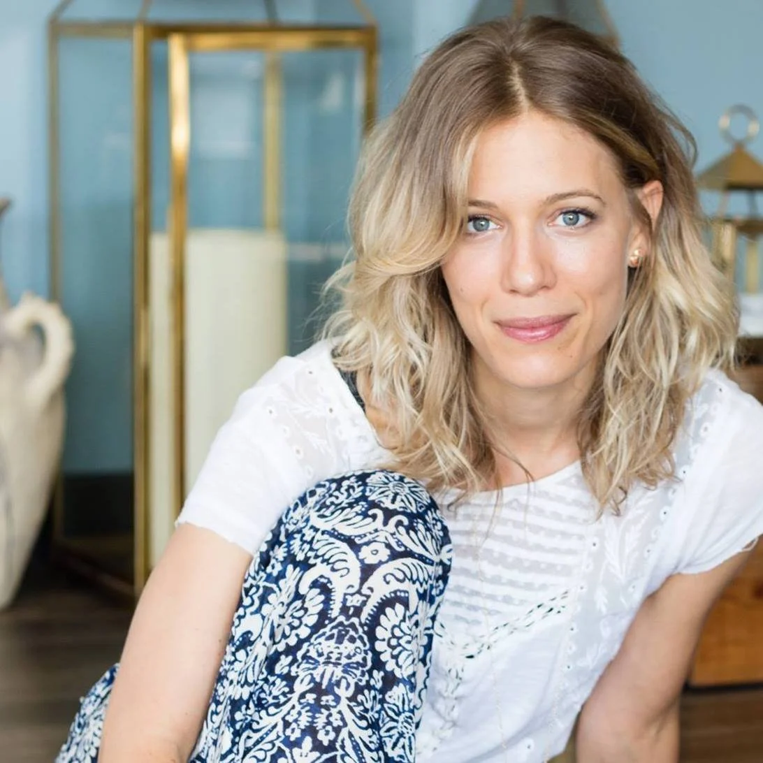 A woman with blonde, wavy hair and blue eyes smiling at the camera, sitting indoors with a blurred background of furniture and decor.