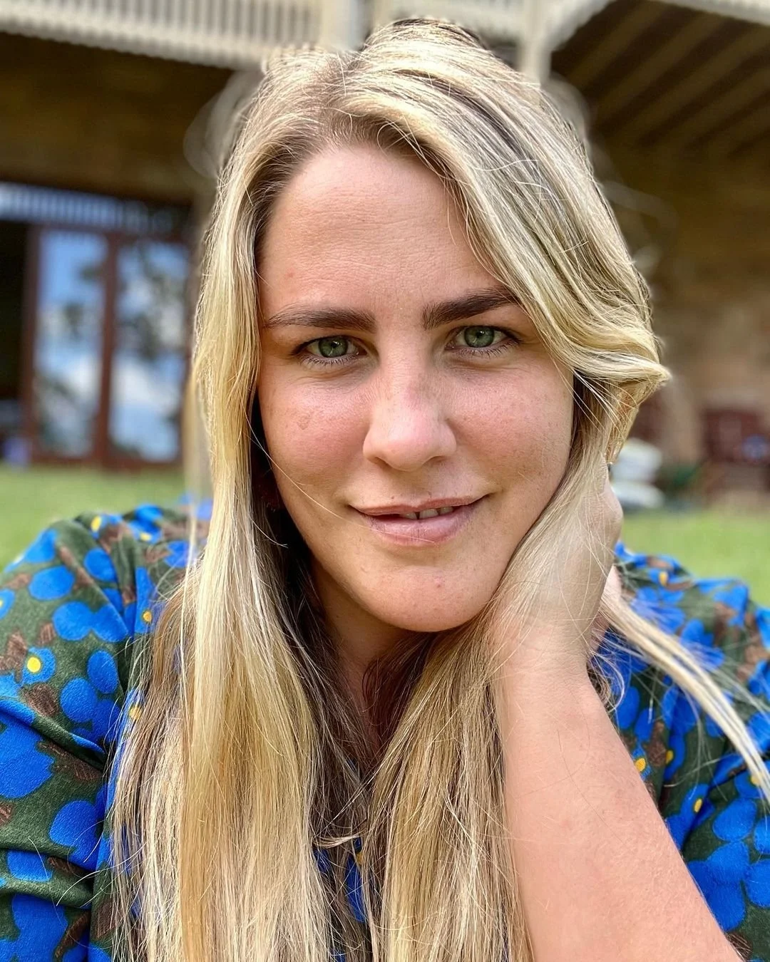 Close-up of a young woman with long blonde hair and green eyes smiling, wearing a blue patterned top, outdoors with a blurred background of a house and garden.