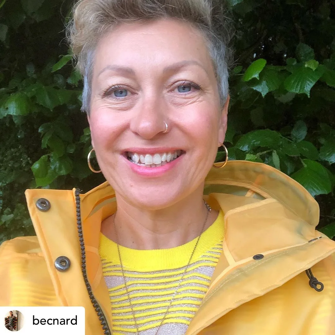 A woman with short gray hair and blue eyes smiling outdoors, wearing gold hoop earrings, a yellow striped shirt, and a yellow rain jacket, standing in front of green foliage.
