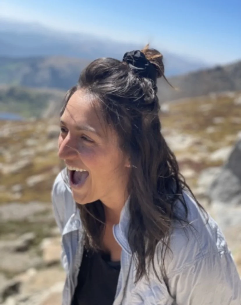 Woman with dark hair tied in a bun, smiling and laughing outdoors in a mountainous landscape.