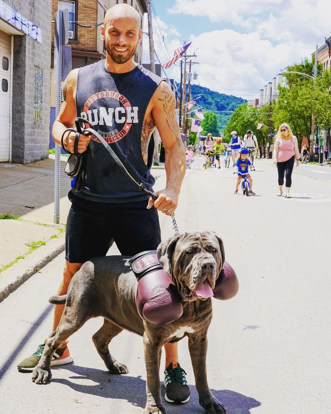 A smiling man wearing a black tank top and shorts stands on a city street holding a leash attached to a large dog. The dog, a brindle mastiff, has boxing gloves around its neck and is panting with its tongue out. In the background, there are pedestrians, including children on bicycles, walking along the street with American flags and greenery visible.