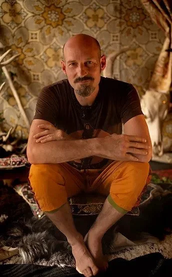 Man sitting cross-legged on a rug with arms crossed, in a room with patterned wallpaper and decorative elements in the background.