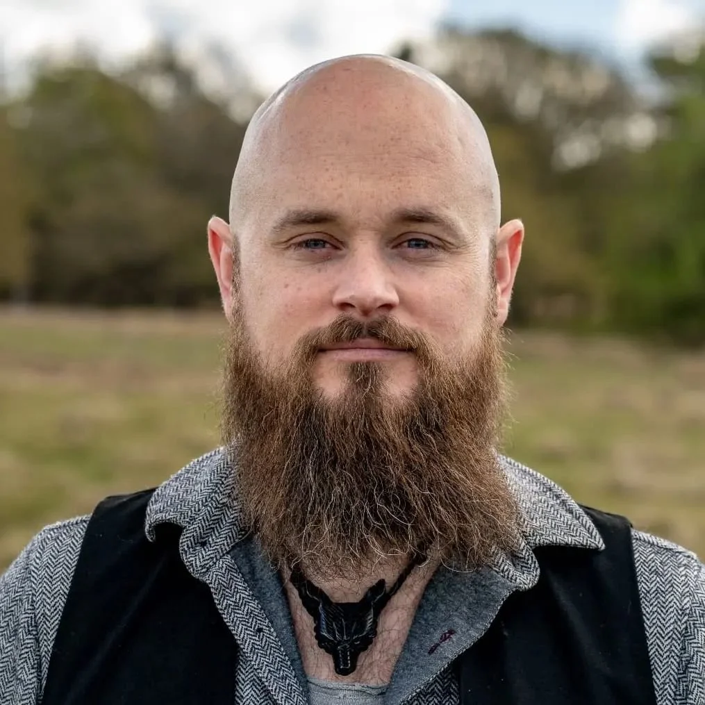 A bald man with a full beard, wearing a gray patterned shirt and a black vest, standing outdoors in a natural setting.
