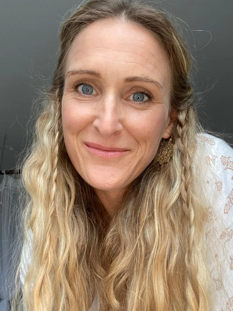 Close-up of a woman with long, wavy blonde hair, blue eyes, and a gentle smile, wearing earrings and a white top with a subtle pattern.