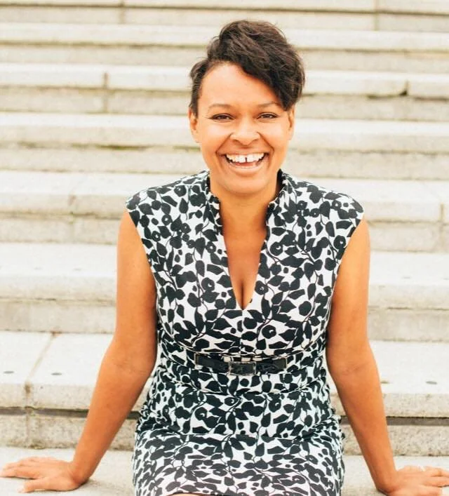 A woman sitting on outdoor steps, smiling, wearing a black and white patterned dress.