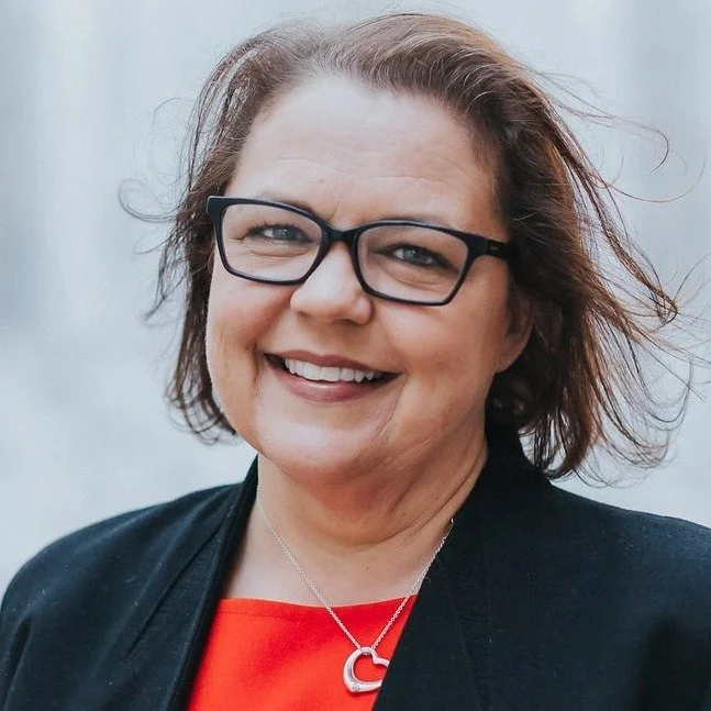 A woman with short brown hair, glasses, and a bright smile, wearing a black blazer over a red top and a silver necklace with a heart-shaped pendant, standing outdoors with a neutral background.