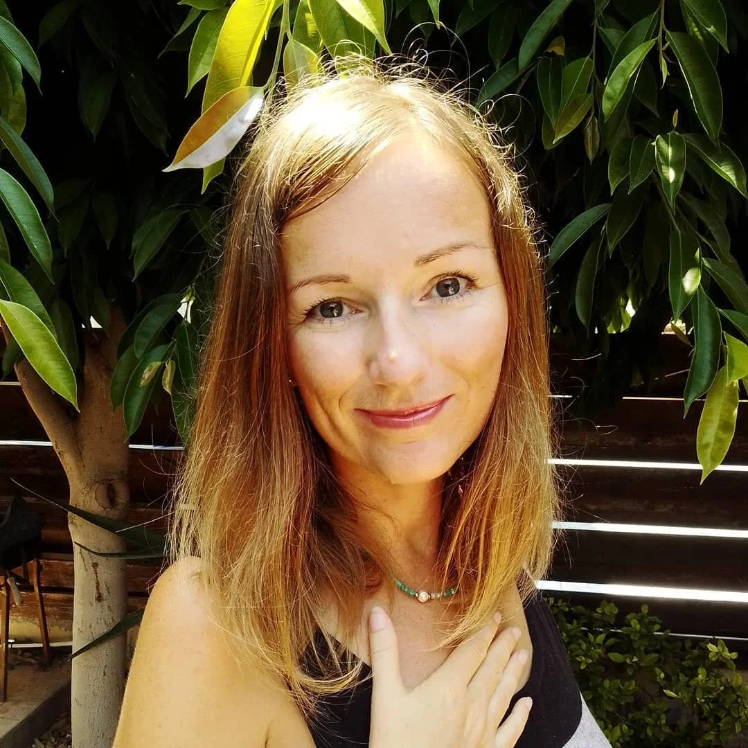 A woman with long red hair and fair skin standing outdoors in front of green leafy trees, smiling at the camera.