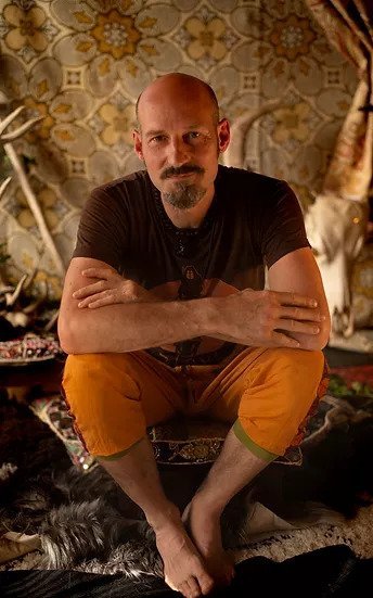 A man with a shaved head and goatee, wearing a black t-shirt and yellow pants, is sitting cross-legged on the floor with arms crossed in front of a decorative, patterned background.
