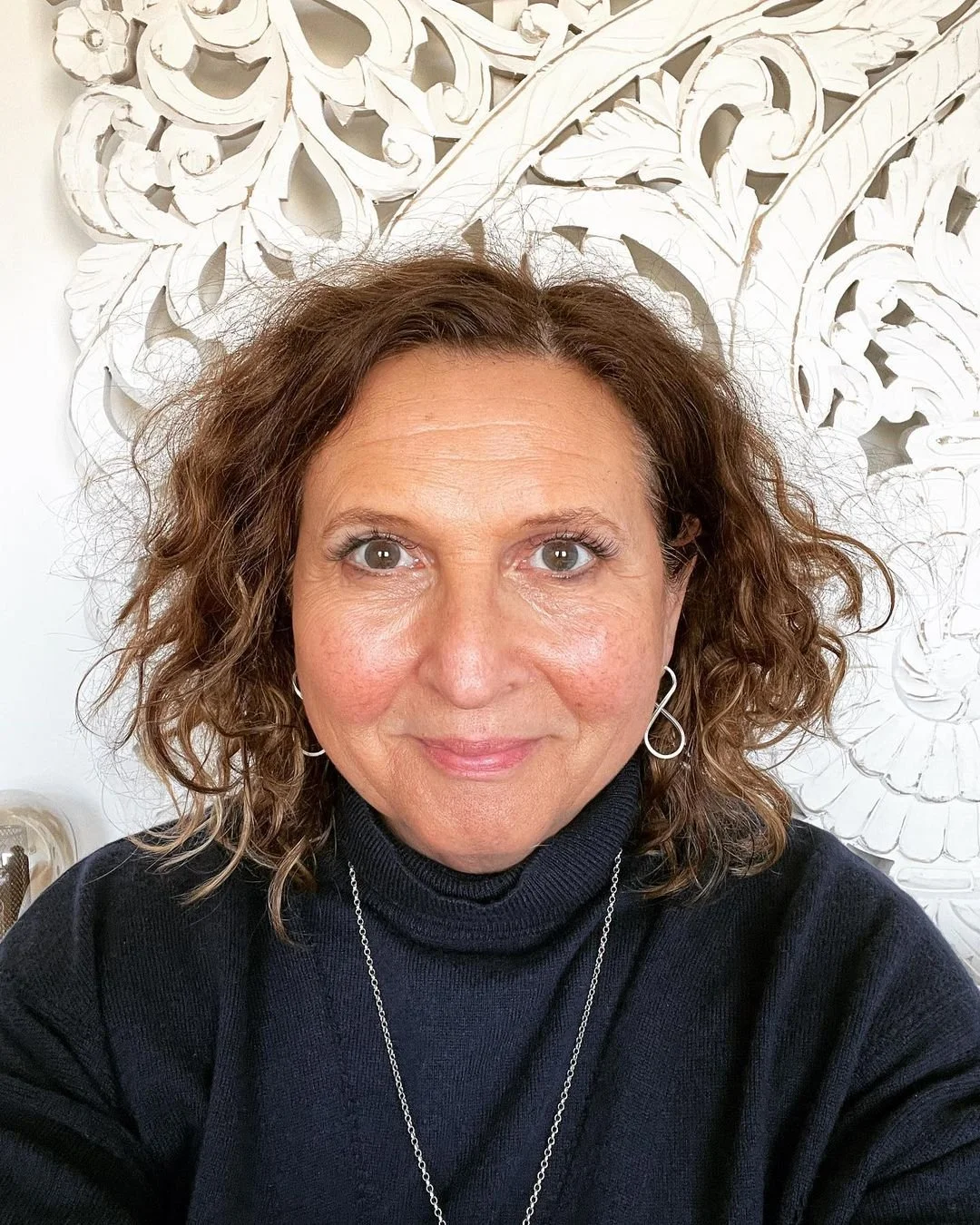 A woman with wavy brown hair and earrings, smiling, wearing a black turtleneck and a necklace, sitting in front of a white decorative wall.