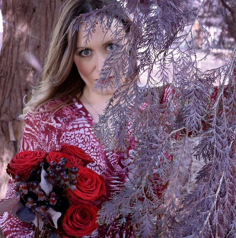 A woman with wavy blonde hair and piercing blue eyes, partially hidden behind purple-tinted leaves, holds a bouquet of red roses and dark berries in a natural, wooded setting.