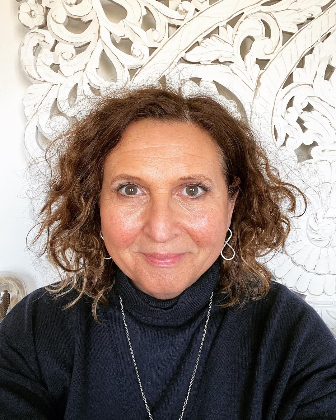 A woman with curly brown hair, wearing a black turtleneck sweater and silver earrings, taking a selfie. Behind her is a white decorative wall with intricate floral patterns.