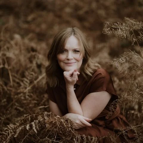 A woman with shoulder-length brown hair smiling, resting her chin on her right hand, sitting outdoors among brown foliage and plants.