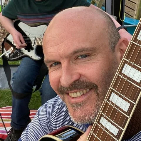 A smiling man holding an acoustic guitar, with another person playing an electric guitar in the background.