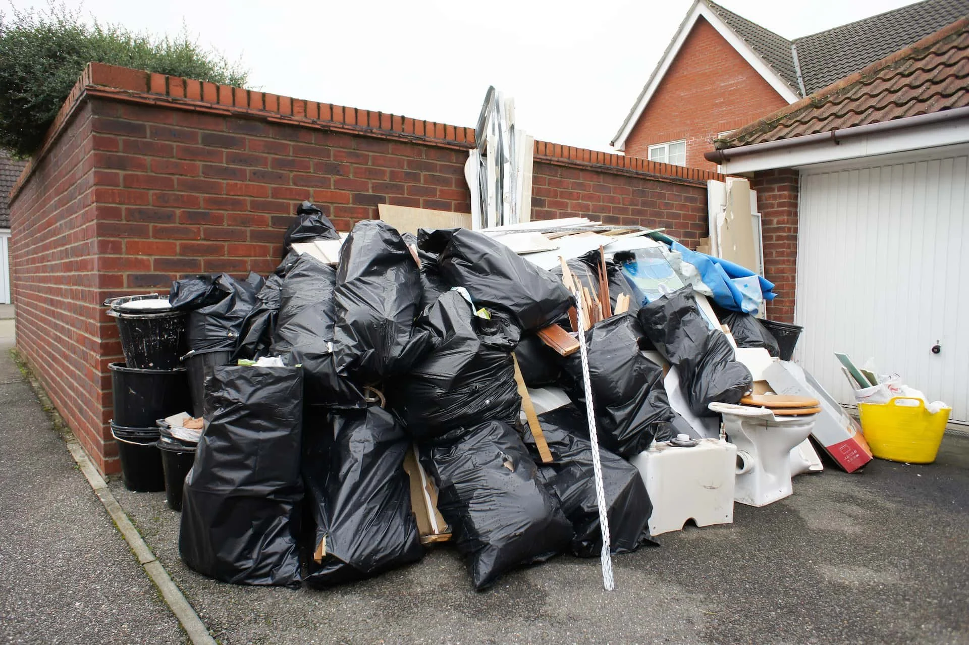 Large pile of household trash bags, furniture, and debris outside against a brick wall.