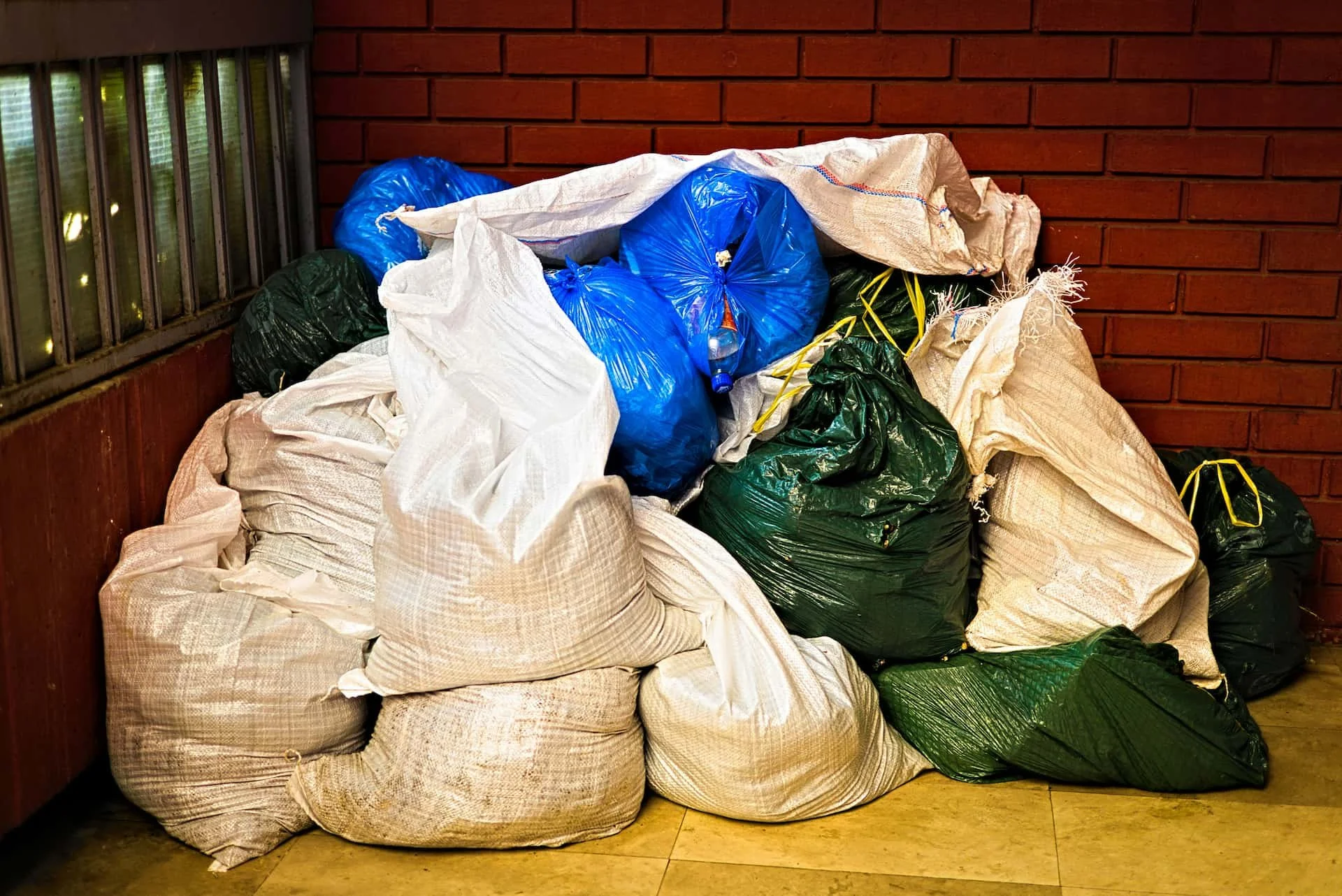 Piles of garbage bags in various colors including blue, green, and white, stacked against a red brick wall with a window on the side.