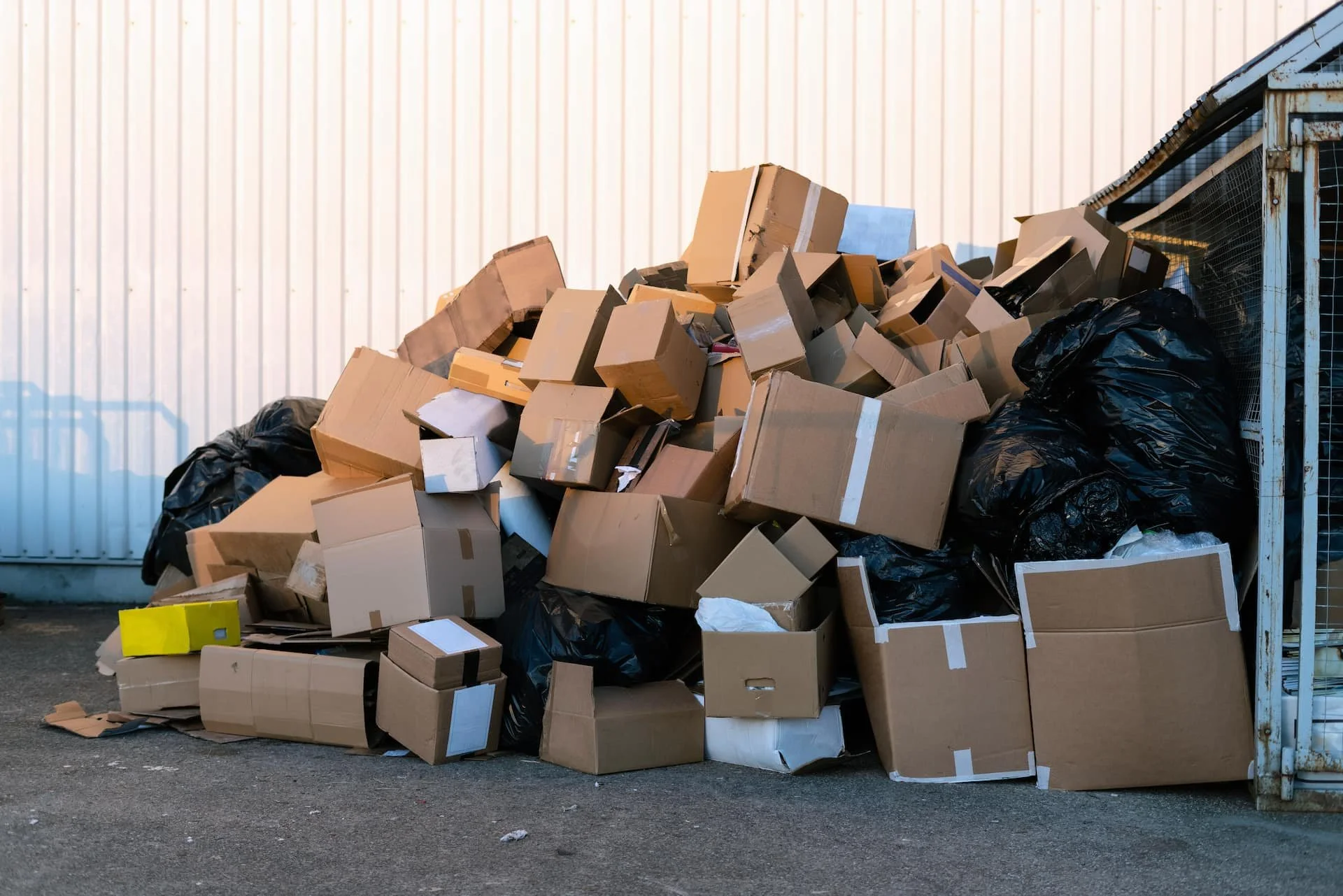 Piles of cardboard boxes and black trash bags outside against a white wall.