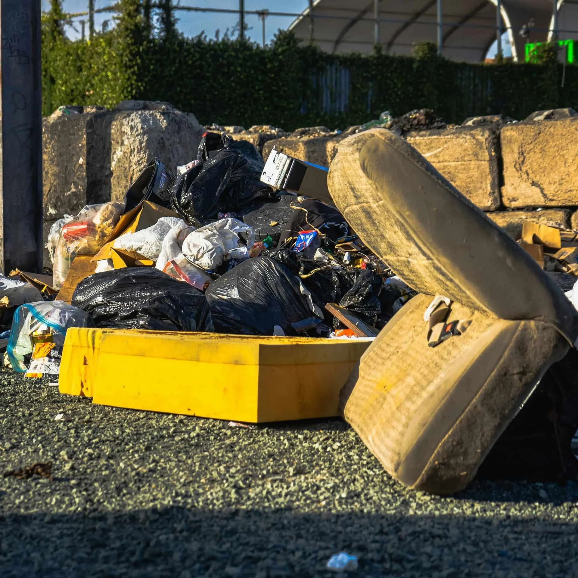 Pile of trash and debris with black plastic bags and a yellow bin, scattered on asphalt near a stone wall and bushes.