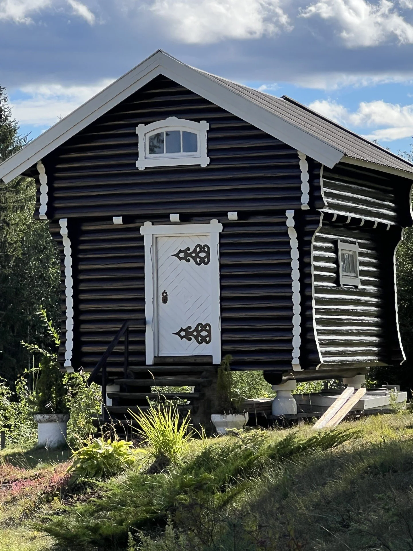 En liten, svart og hvit trehytte med trapp foran, omgitt av grønt gress og planter, under en blå himmel med skyer.