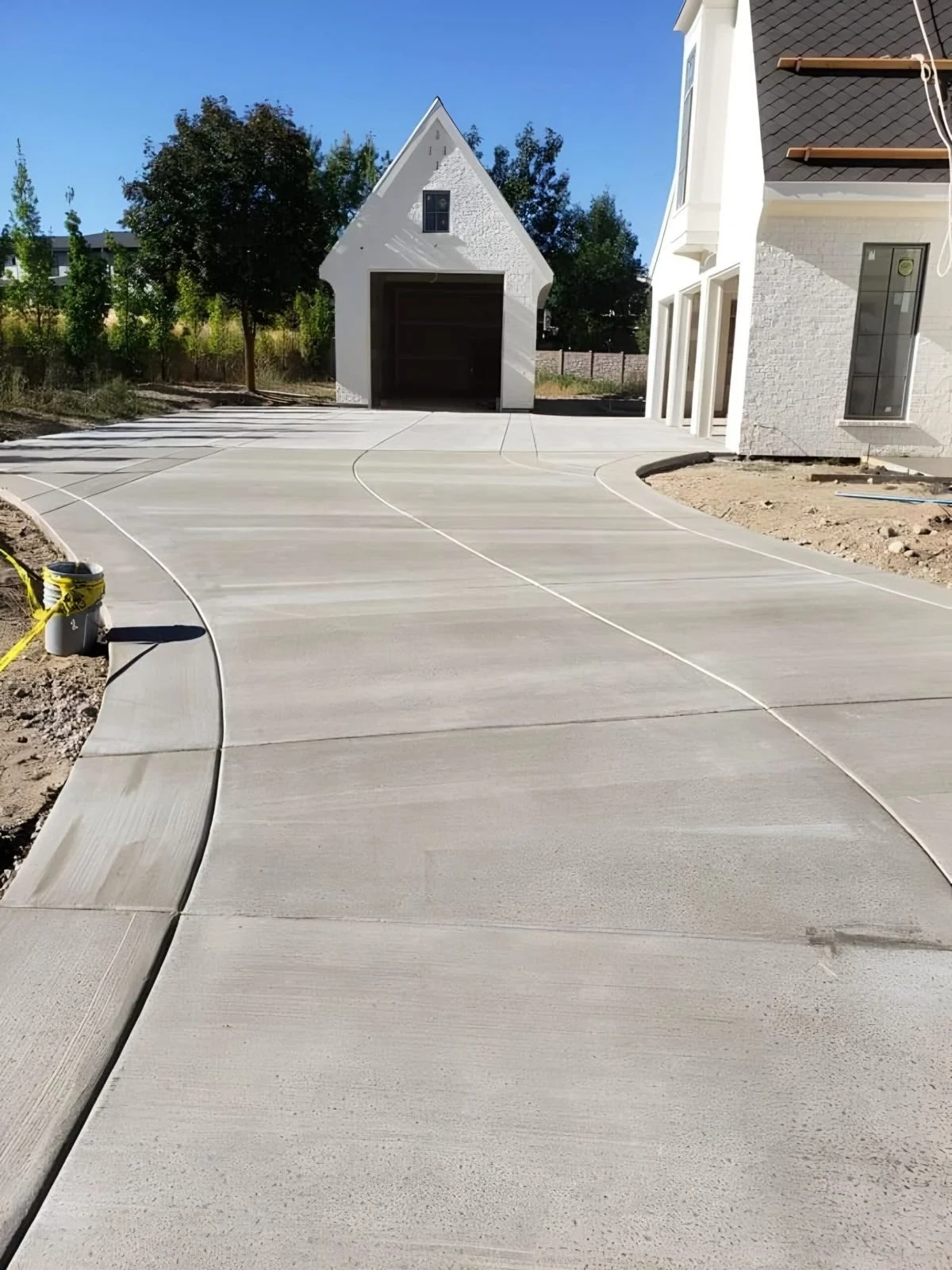 Newly poured concrete driveway in front of a white house with a garage and a small building in the background, with some trees and clear blue sky.