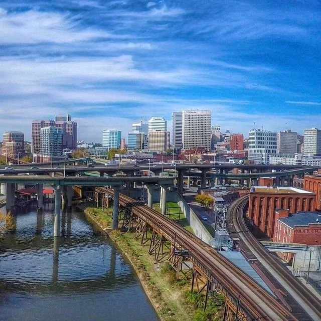 City skyline with tall buildings, elevated train tracks over a river, and a bright blue sky with scattered clouds.