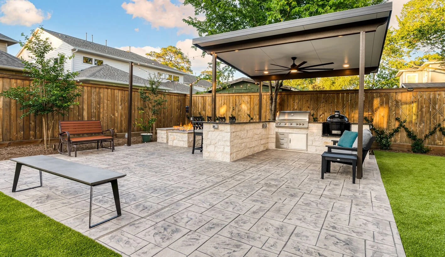 Backyard patio with outdoor kitchen, seating areas, and a fire pit, surrounded by a wooden fence and greenery.