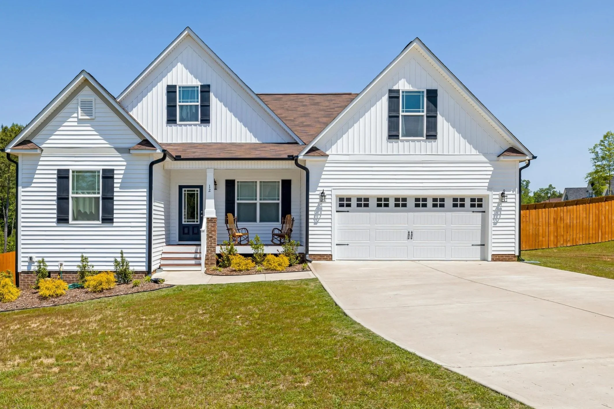 A white two-story house with black shutters, a brown shingled roof, and a two-car garage. There's a small porch with two chairs and a garden with shrubs and yellow flowers in front.