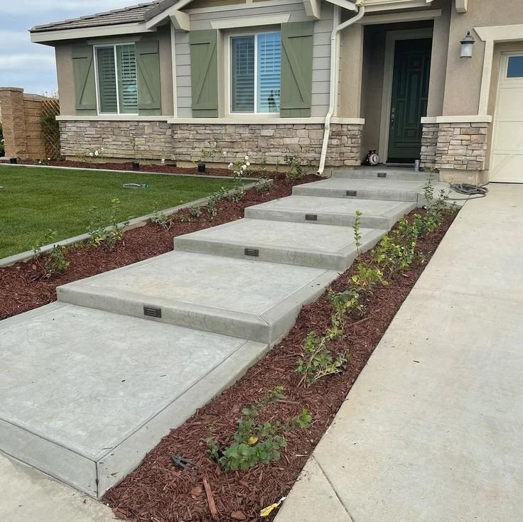 Concrete walkway with steps leading up to a house entrance, flower bed with small plants and mulch on each side, house with green shutters and stone accent wall.