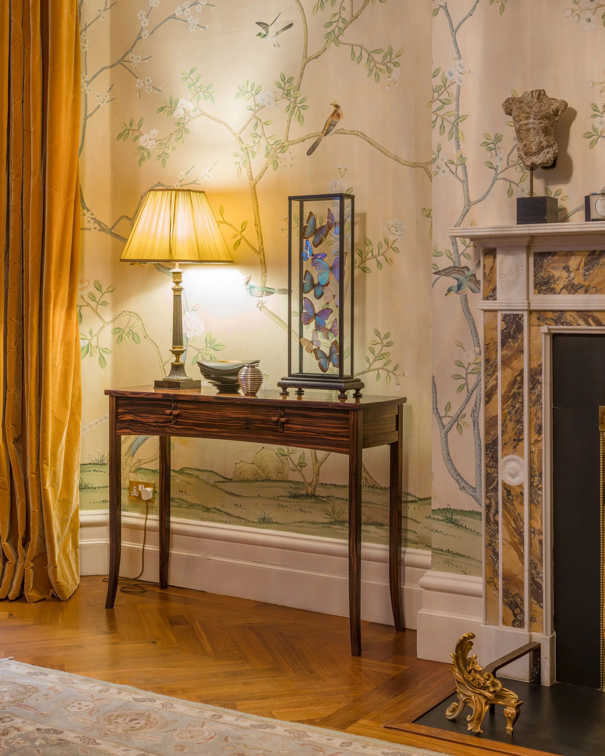 An interior scene with a wooden side table, a lit table lamp, a decorative glass display with butterflies, and a painted wallpaper with birds and branches. A yellow curtain is visible on the left, and part of a fireplace with marble trim is on the ri
